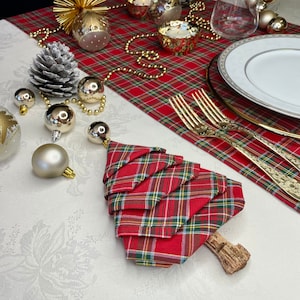 May include: A red and green tartan napkin folded into the shape of a Christmas tree. The napkin is sitting on a white tablecloth with gold baubles and a pine cone.