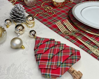 May include: A red and green tartan napkin folded into the shape of a Christmas tree. The napkin is sitting on a white tablecloth with gold baubles and a pine cone.