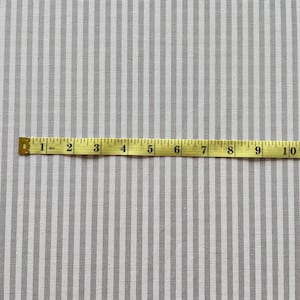 Puede incluir: Un primer plano de una tela blanca con finas rayas grises. Una cinta m&eacute;trica amarilla se coloca sobre la tela, mostrando medidas de 1 a 10 pulgadas.