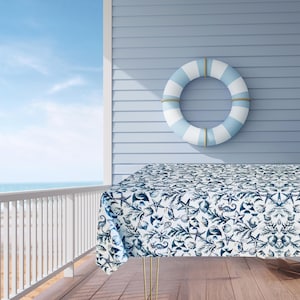 May include: A white tablecloth with a blue and white nautical pattern featuring seashells, starfish, and seaweed. The tablecloth is laid on a table on a wooden deck with a white railing and a blue and white striped life preserver hanging on the wall behind it.