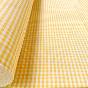 Può includere: Un rotolo di tessuto a quadretti giallo e bianco. Il tessuto presenta un classico motivo a quadretti. Il rotolo è parzialmente srotolato, rivelando la trama e il colore del tessuto. Il motivo è uniforme su tutta la superficie visibile.