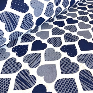 Puede incluir: Tela blanca con un patrón de corazones azul marino y gris. Los corazones presentan varios diseños, incluyendo rayas, lunares y pata de gallo. La tela está doblada en la imagen, mostrando la continuidad del patrón.