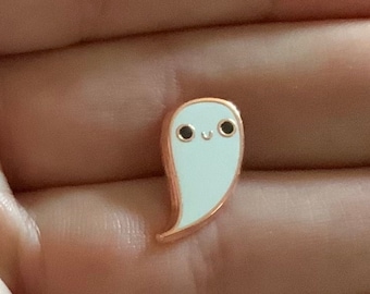 May include: A white enamel pin shaped like a ghost with a smiling face. The pin has a rose gold finish.