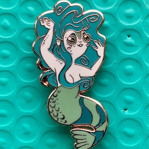 Pode incluir: Um alfinete esmaltado de sereia verde e turquesa com contorno em ouro rosa. A sereia tem cabelo turquesa, tronco branco e cauda verde e azul. O alfinete está em um fundo turquesa.