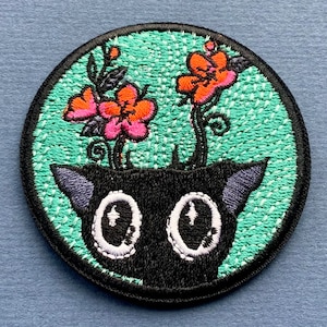 Pode incluir: Etiqueta bordada com um gato preto de olhos grandes e um arranjo floral de flores cor-de-rosa e laranja sobre um fundo turquesa.