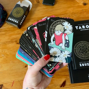 Pu&ograve; includere: Un mazzo di carte dei tarocchi con illustrazioni colorate su sfondi neri. Le carte sono disposte a ventaglio in una mano, rivelando la carta "Il Matto" con una figura infantile in un vestito rosso. Le carte sono etichettate "Next Gen Tarot by Zsuzsa".
