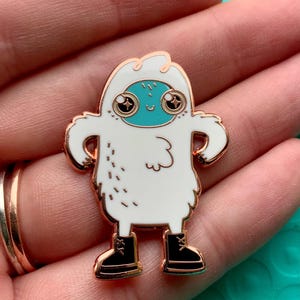 Pode incluir: Um alfinete de esmalte branco com um yeti de desenho animado com um rosto turquesa, olhos grandes e botas pretas. O alfinete tem um contorno dourado e é segurado em uma mão.