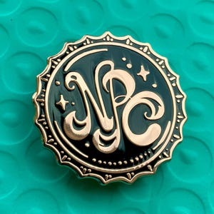 Puede incluir: Un pin redondo de esmalte negro y dorado con las letras "NPC" en una fuente estilizada. El pin tiene un borde dorado con un borde festoneado y pequeños detalles de estrellas. El fondo es turquesa.