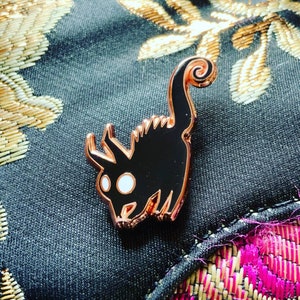 May include: A black and rose gold enamel pin of a stylized creature with a curled tail and a white eye.