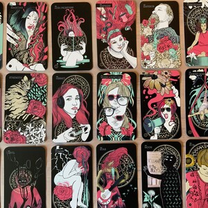 Pu&ograve; includere: Un set di 22 carte dei tarocchi con illustrazioni colorate che raffigurano persone e simboli. Le carte hanno uno sfondo nero con accenti dorati. I nomi delle carte sono stampati in lettere dorate nella parte superiore di ogni carta. Le carte sono disposte in tre file di sette carte.