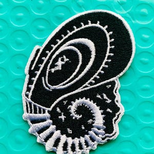 May include: Black and white embroidered patch featuring a stylized seashell design. The patch has a detailed, textured appearance with intricate stitching. The seashell design is set against a teal background.