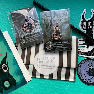 May include: A set of three black and white enamel pins featuring a cartoon monster with large eyes and horns. The pins are packaged in clear plastic bags with black and white striped paper backing. The pins are labeled "isolation monster" and "anxiety".