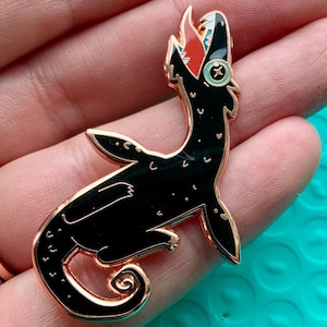 May include: A black enamel pin in the shape of a sea creature with a gold outline. The creature has a red mouth, a green eye, and white accents. The pin is on a light blue surface.