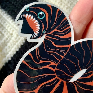 May include: A black and orange sticker of a cartoon sea creature with a large mouth and two eyes. The creature is curled up in a circle.