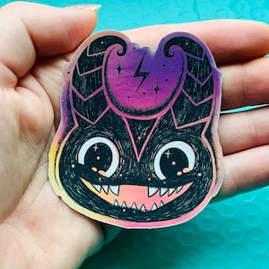 Puede incluir: Una pegatina holográfica de un monstruo de dibujos animados con un fondo degradado morado, rosa y amarillo. El monstruo tiene ojos negros, una amplia sonrisa y dos cuernos. Hay un rayo y estrellas en la cabeza del monstruo.