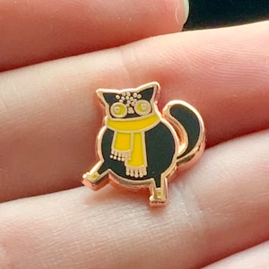 Könnte beinhalten: Eine schwarze Emaille-Anstecknadel mit einem Cartoon-Katzenmotiv, das einen gelben Schal trägt. Die Anstecknadel hat eine roségoldene Oberfläche.