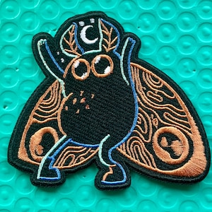 Op de afbeelding: Geborduurde patch met een gestileerde zwarte mot met grote ogen, een halve maan en ingewikkelde vleugelpatronen in oranje en bruin. De mot heeft blauwe en witte accenten, wat een grillig ontwerp creëert. Geschikt voor jassen, tassen of andere stoffen.