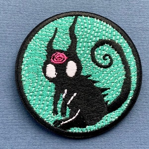 May include: A black embroidered patch with a stylized creature with a pink flower on its head. The creature is sitting on a green background with a spiral design.