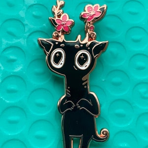 Puede incluir: Pin de esmalte negro con una criatura caprichosa con flores rosas en la cabeza. La criatura tiene ojos grandes y una cola enroscada. El pin tiene un acabado dorado.