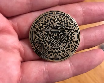 Può includere: Uno spillo in metallo dorato con un design in smalto nero. Il design presenta una stella a cinque punte con una luna crescente e un cerchio al suo interno. La stella è circondata da un cerchio di linee ondulate.