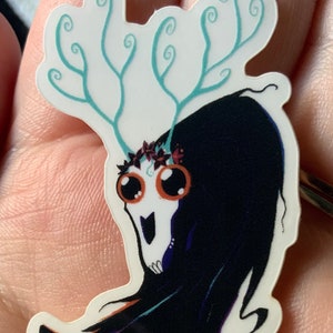 Op de afbeelding: Een sticker met een zwart-witte illustratie van een schedel met grote ogen en een kroon van geweien. De geweien zijn lichtblauw en hebben een draaiend ontwerp.