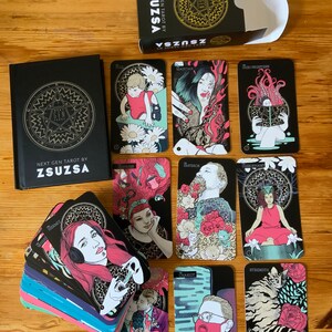 Pu&ograve; includere: Un mazzo di carte dei tarocchi con bordi neri e dorati e illustrazioni colorate. Le carte sono disposte a ventaglio su una superficie di legno. La scatola delle carte &egrave; nera con lettere dorate che recitano "Next Gen Tarot by Zsuzsa".