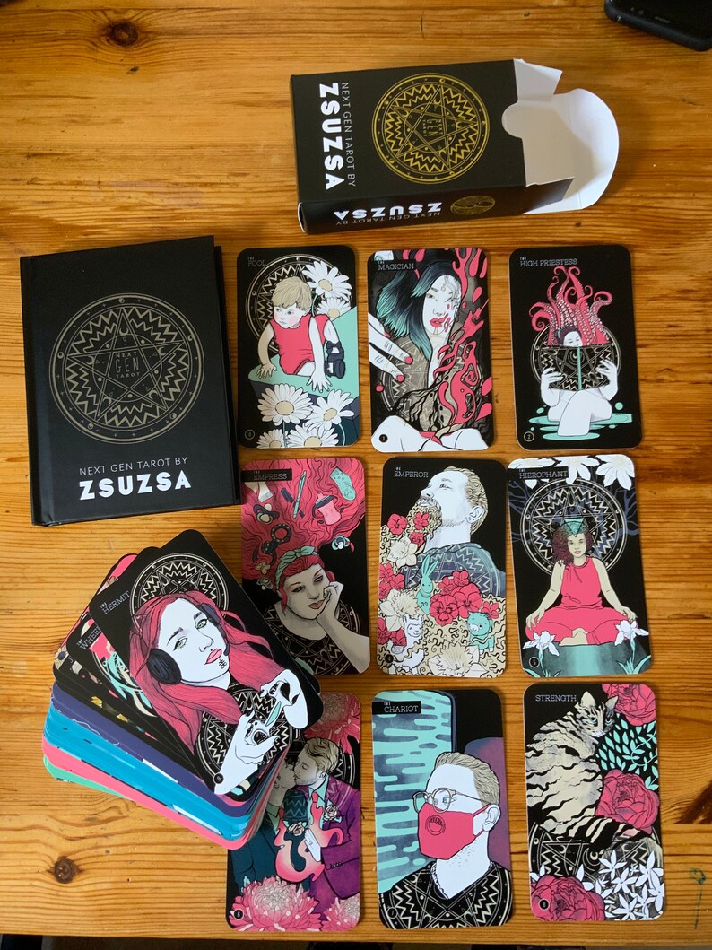 Pu&ograve; includere: Un mazzo di carte dei tarocchi con bordi neri e dorati e illustrazioni colorate. Le carte sono disposte a ventaglio su una superficie di legno. La scatola delle carte &egrave; nera con lettere dorate che recitano "Next Gen Tarot by Zsuzsa".