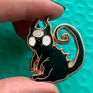 Könnte beinhalten: Eine schwarze Emaille-Anstecknadel mit roségoldener Oberfläche, die eine stilisierte Katze mit einem wirbelnden Schwanz und drei Augen darstellt. Die Augen der Katze sind weiß mit schwarzen Pupillen und das dritte Auge ist eine Spirale.