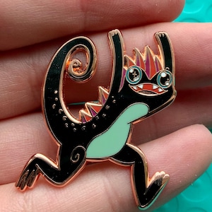 May include: A black and gold enamel pin featuring a cartoon lizard with a light green belly, red and purple spikes, and a curled tail. The lizard is in a playful pose with its arms raised. The pin has a gold outline.