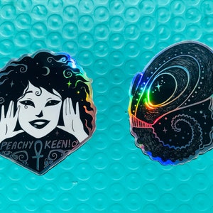May include: Two holographic stickers, one featuring a black and white illustration of a woman with curly hair and the text "Peachy Keen!" and the other featuring a black and white illustration of a creature with a spiral design.