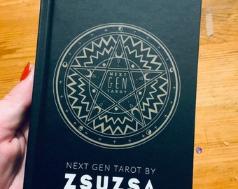 Può includere: Un libro nero con un disegno a stella dorata e il testo "Next Gen Tarot by Zsuzsa".