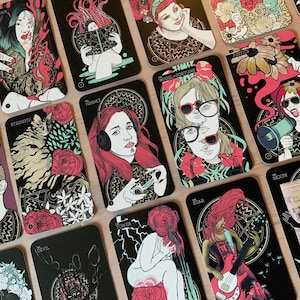 Puede incluir: Un conjunto de cartas de tarot con fondos negros e ilustraciones coloridas. Cada carta presenta obras de arte únicas, incluyendo retratos, flores e imágenes simbólicas. Las cartas están dispuestas sobre una superficie de madera clara.