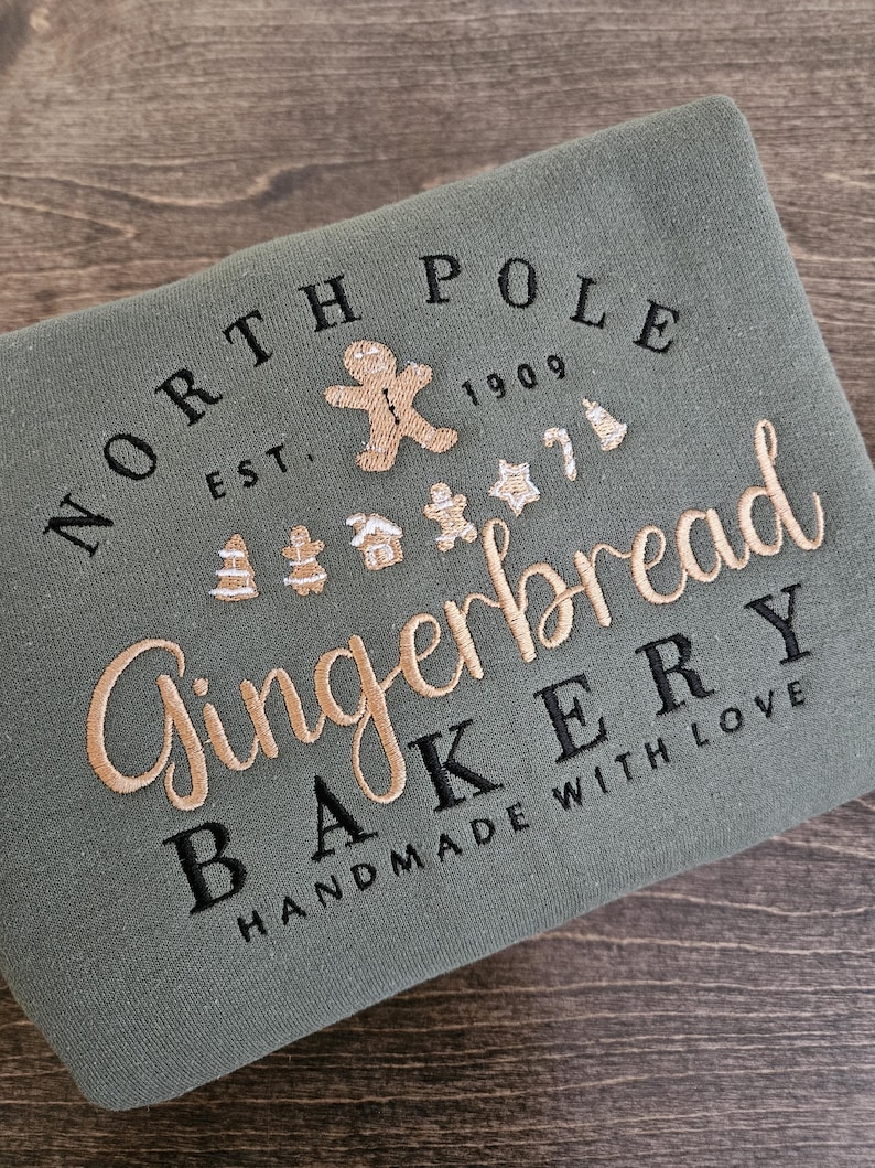May include: Green sweatshirt with embroidered text that reads "North Pole Gingerbread Bakery Est. 1909 Handmade with Love". The text is surrounded by gingerbread man and other holiday-themed imagery.