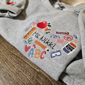 May include: Grey sweatshirt with embroidered school supplies, including an apple, a pencil, a ruler, a crayon, scissors, a glue stick, and the letters ABC. The text "Ms. Nikki" is also embroidered on the sweatshirt.