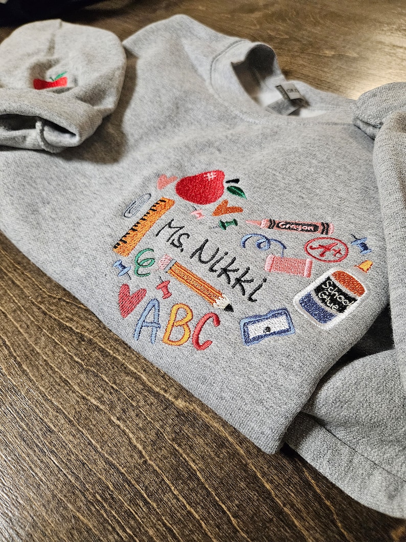May include: Grey sweatshirt with embroidered school supplies, including an apple, a pencil, a ruler, a crayon, scissors, a glue stick, and the letters ABC. The text "Ms. Nikki" is also embroidered on the sweatshirt.