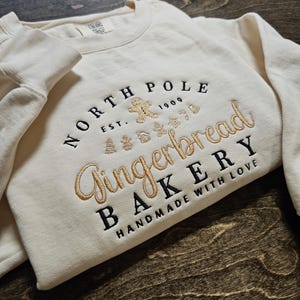 May include: A cream-colored sweatshirt with a gold embroidered design that says "North Pole Gingerbread Bakery Handmade with Love" with a gingerbread man and other Christmas-themed imagery.