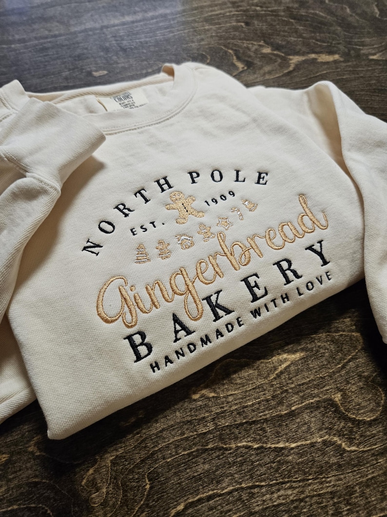 May include: A cream-colored sweatshirt with a gold embroidered design that says "North Pole Gingerbread Bakery Handmade with Love" with a gingerbread man and other Christmas-themed imagery.