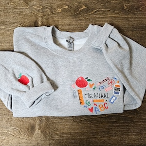 May include: Grey sweatshirt with a red apple and school supplies embroidered on the front. The text "Ms. Nikki" is embroidered below the design. The sweatshirt has a matching grey beanie with a red apple embroidered on it.