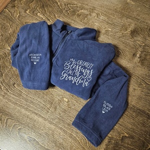 May include: A navy blue fleece jacket with the text "My Greatest Blessings Call Me Grandma" embroidered on the front. The jacket has two pockets on the front and a zipper closure. The text "Jackson, Emilia, Rosie" is embroidered on the left pocket and the text "Elissa, Silas, Levi" is embroidered on the right sleeve.