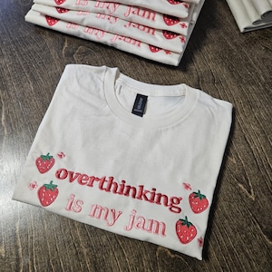 Puede incluir: Camiseta de color crema con la frase bordada "overthinking is my jam" en rojo y rosa. La camiseta presenta fresas bordadas. Se ve una pila de camisetas similares en el fondo.