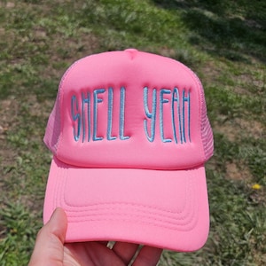 May include: A pink trucker hat with the embroidered text "SHELL YEAH" in light blue. The hat has a mesh back and a curved brim. The hat is held up by a hand.