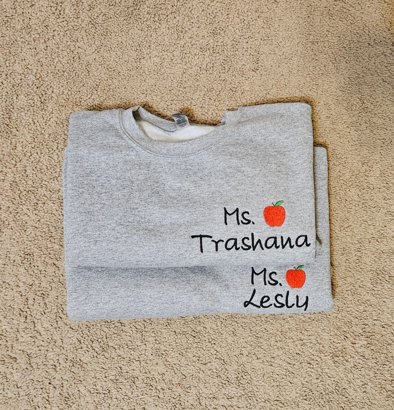 May include: Grey sweatshirt with the text "Ms. Trashana" and a red apple graphic, and the text "Ms. Lesly" and a red apple graphic.
