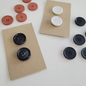 May include: Two sets of buttons on brown card stock. One set is black, the other is white. A third set of buttons is orange and scattered on a white surface.