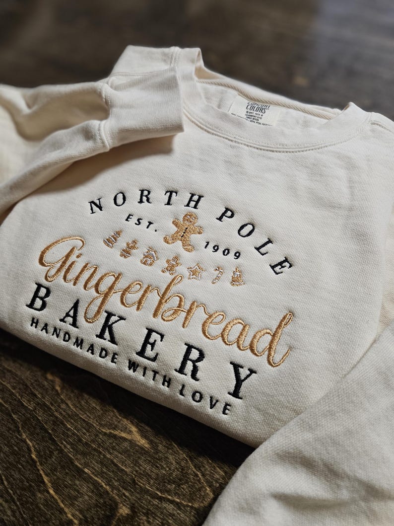May include: A white sweatshirt with the text "North Pole Gingerbread Bakery Handmade with Love" in black and gold embroidery. The text is in a circular design with a gingerbread man in the center.