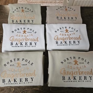 May include: Six beige and white sweatshirts with the text "North Pole Gingerbread Bakery Handmade with Love" embroidered on the front. The text is in a gold and brown color scheme.