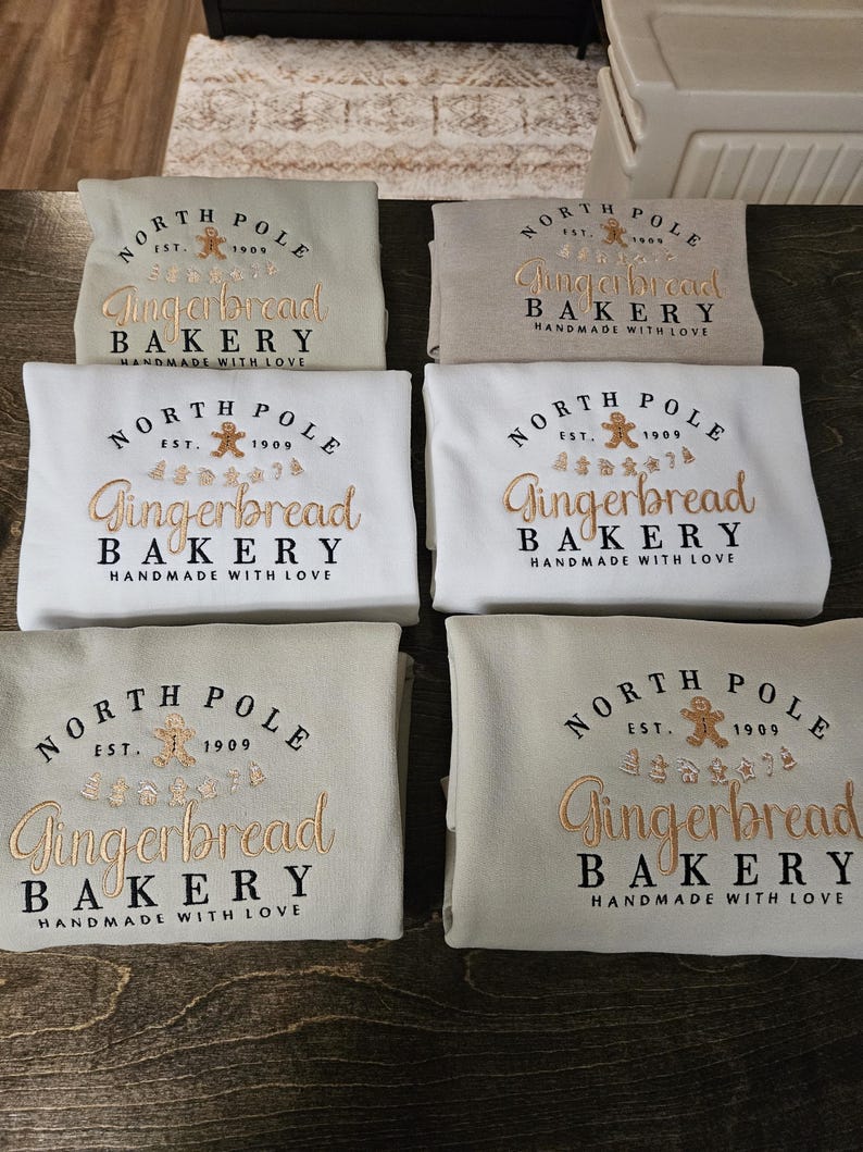 May include: Six beige and white sweatshirts with the text "North Pole Gingerbread Bakery Handmade with Love" embroidered on the front. The text is in a gold and brown color scheme.