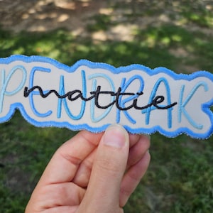 以下が含まれることがあります： 「PENDRAK mattie」という名前がスタイリッシュなフォントで刺繍された、水色と白のパッチ。名前は黒の筆記体で、水色のブロック体の文字の上に書かれており、青い縁取りがされています。