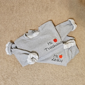 May include: Two grey sweatshirts with red apple embroidery and the text "Ms. Trashana" and "Ms. Lesly" on them.