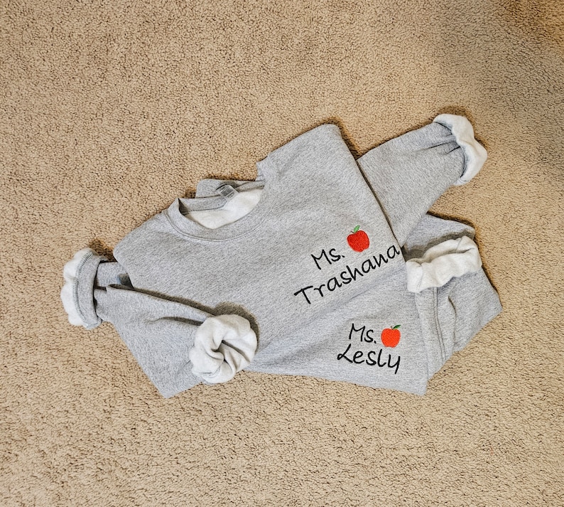 May include: Two grey sweatshirts with red apple embroidery and the text "Ms. Trashana" and "Ms. Lesly" on them.