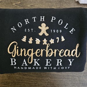 May include: Black sweatshirt with white embroidered text that reads "North Pole Gingerbread Bakery Est. 1909 Handmade with Love". The text is surrounded by a gingerbread man and gingerbread cookies.